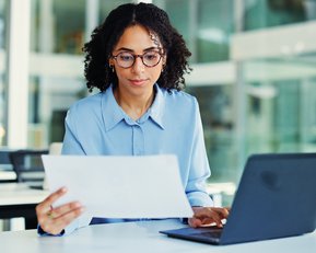 Eine Frau mit lockigem Haar und Brille sitzt an einem Schreibtisch, schaut auf ein Dokument und arbeitet am Laptop.