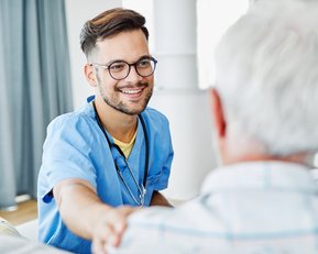 Ein junger Arzt in blauer Arbeitskleidung lächelt und bietet einem älteren Mann, der ihm zugewandt ist, die Hand an. Der Arzt trägt ein Stethoskop um den Hals und befindet sich in einer hellen, freundlichen Umgebung.