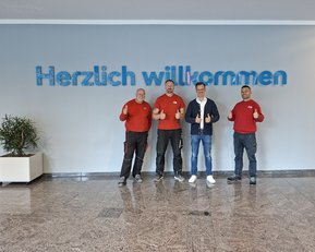 Vier Männer in roten Shirts stehen vor einer Wand mit der Aufschrift "Herzlich willkommen" und zeigen Daumen hoch.