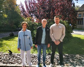 Drei Personen stehen im Freien vor einem grünen Hintergrund mit Bäumen und Sträuchern. Sie tragen legere Kleidung und lächeln.