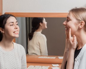 Zwei Frauen stehen vor einem Spiegel. Eine Frau lächelt und zeigt ihre Zähne, während die andere Frau mit einem Finger auf ihr Gesicht zeigt. Im Hintergrund sind einige Notizen sichtbar.