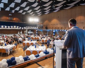 Jubiläum Waldbronn 50 Jahre Ein Redner steht an einem Podium und spricht vor einem Publikum in einem festlich dekorierten Saal mit Tischen und Ballons.