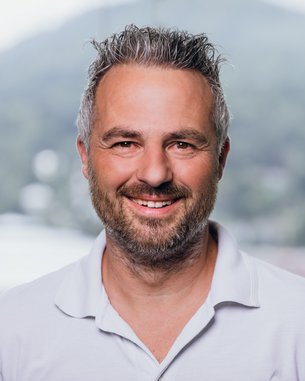 Ein Mann mit grauen Haaren und Bart lächelt in die Kamera. Er trägt ein weißes Polohemd und steht vor einer unscharfen, grünen Landschaft.