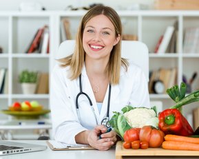 Gesunde Ernährung Eine lächelnde Ärztin in einem weißen Kittel sitzt an einem Tisch mit frischem Gemüse und Obst, während ein Laptop vor ihr steht.