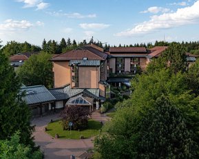 Blick Richtung Haupteingang Gesundheitszentrum Dobel Ein mehrstöckiges Gebäude mit einem modernen Design, umgeben von Bäumen und Grünflächen. Das Gebäude hat ein schräges Dach und große Fenster, die viel Licht hereinlassen.