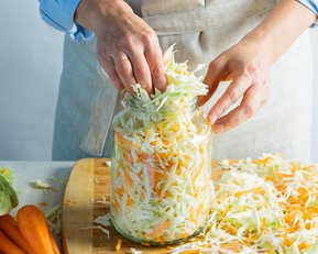 Hände fügen gehobelte Karotten und Kohl in ein großes Glas ein. Um das Glas herum liegen frische Karotten und geschnittenes Gemüse auf einem Holzbrett. Die Person trägt eine helle Schürze.