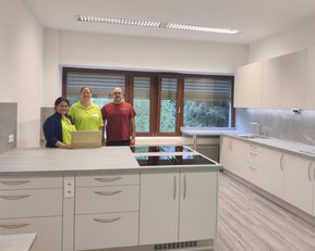 Drei Personen stehen in einer modernen, hellen Küche mit weißen Schränken und einer Kochinsel. Fenster bieten Blick nach draußen.